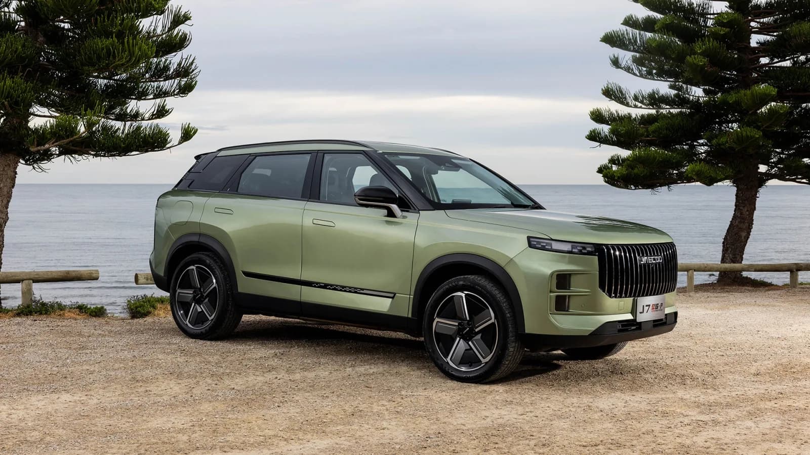 Jaecoo J7 SHS-P in pale green, front three-quarter view, parked on gravel by the sea between two pine trees