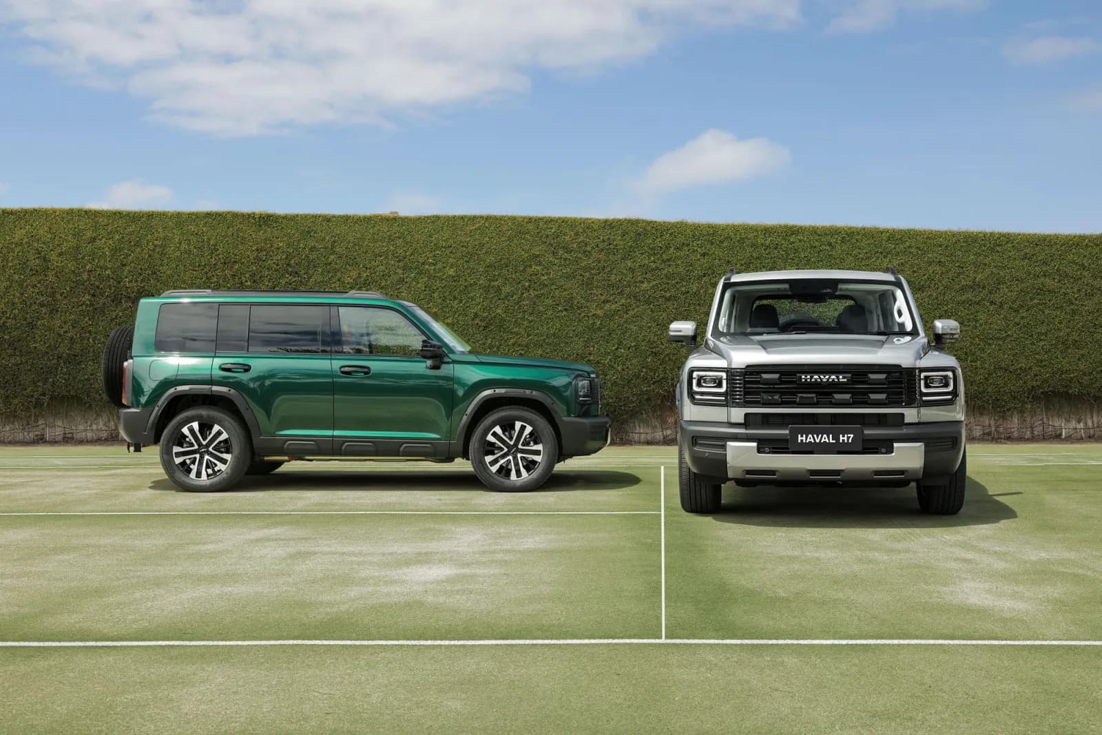 Two new GWM Haval H7 SUVs on a grass tennis court, a green one side-on and a silver one front-on, in front of a tall hedge