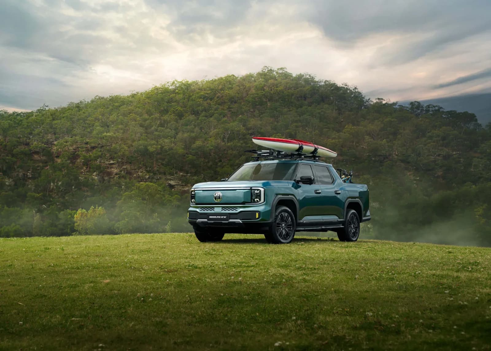 Teal MGU9 EV electric dual-cab ute parked on grass in front of a forested hill, with a kayak on roof racks