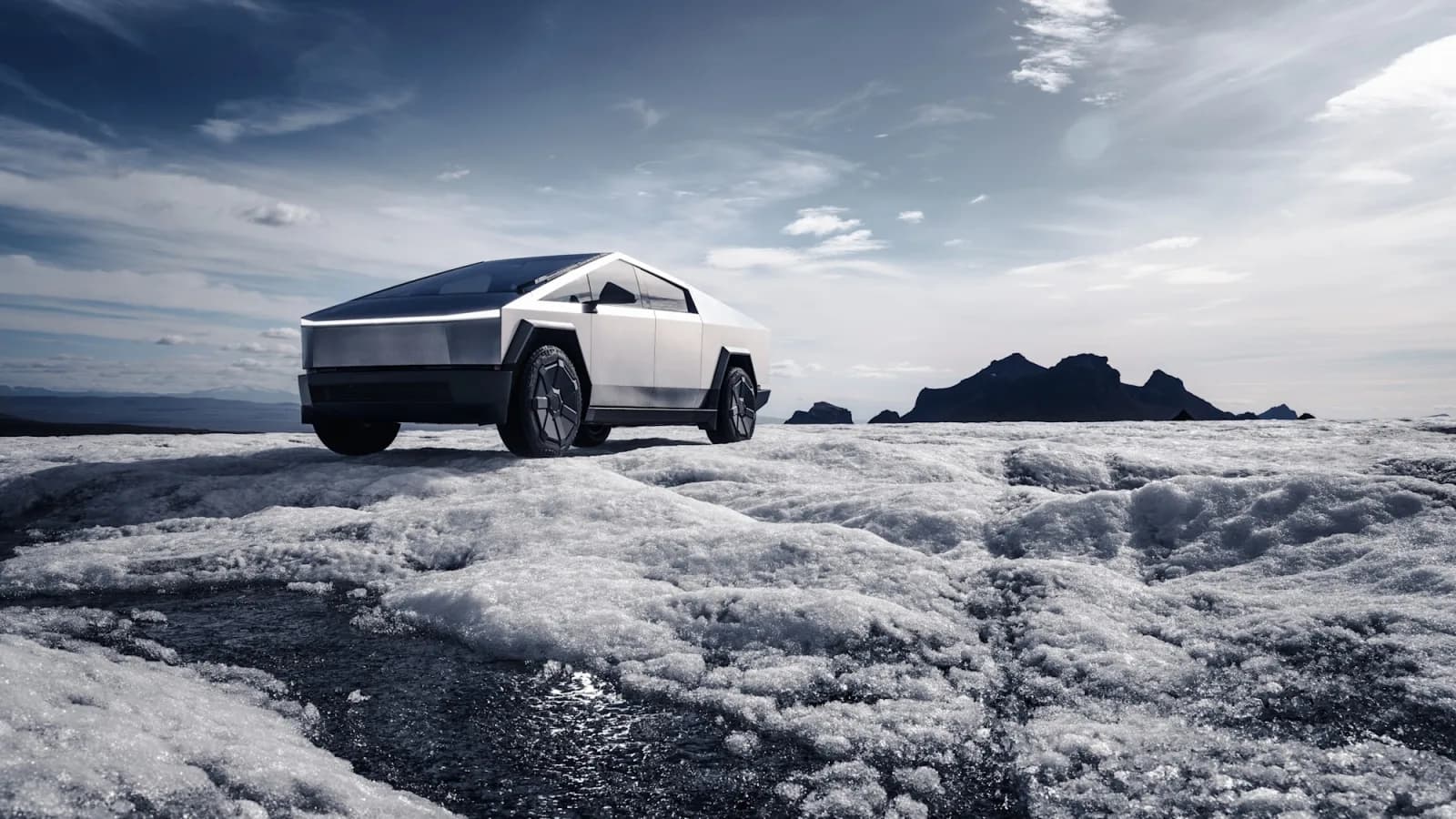 Stainless steel Tesla Cybertruck with its front light bar on, parked on a glacier under a cloudy sky